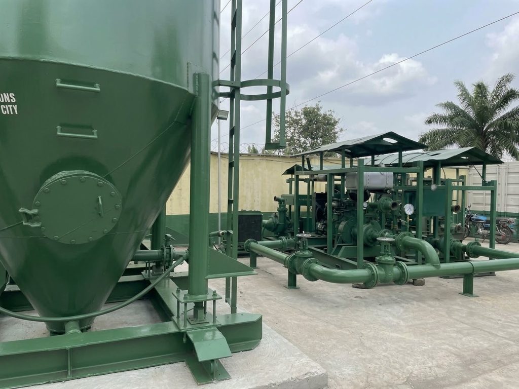 Green cone-bottom bulk silo beside covered pump skids and pipework